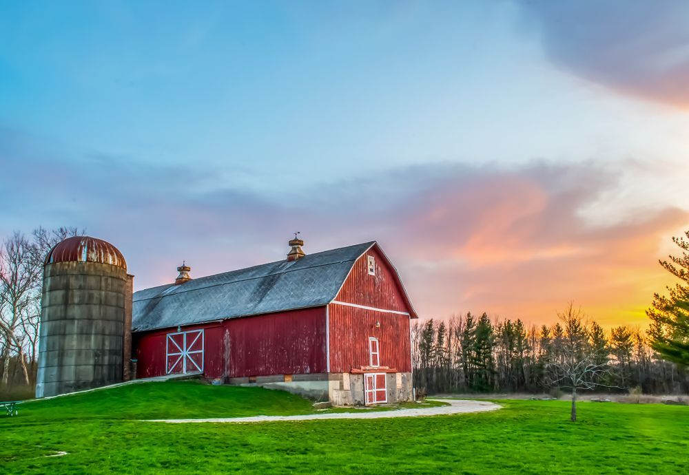 Red barn and silo on green grass at sunset with colorful sky.