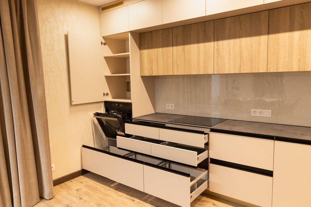 Modern kitchen with light wood and cream cabinetry, open drawers, and built-in appliances.