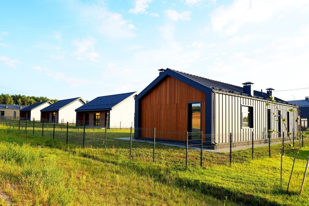 Row of modern houses with brown and white siding and dark roofs, behind a small fence, in a grassy field.