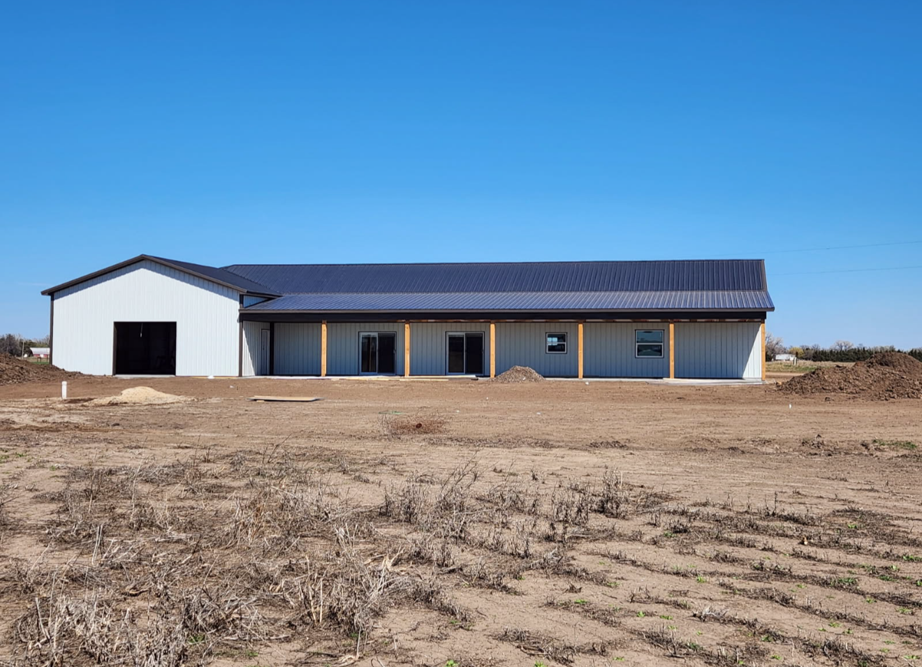 Steel-framed building with a dark roof and a covered porch on a dirt lot under a blue sky.