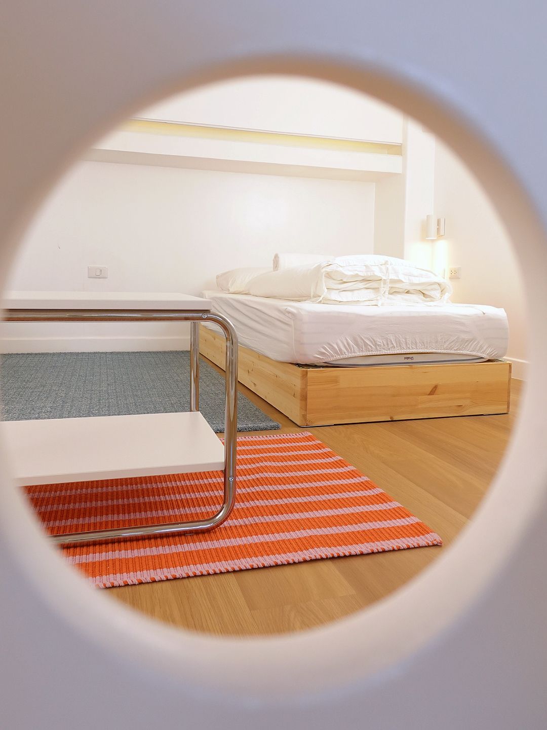 Bedroom scene seen through a porthole; bed, chair, rug. Orange and white striped rug in the foreground.