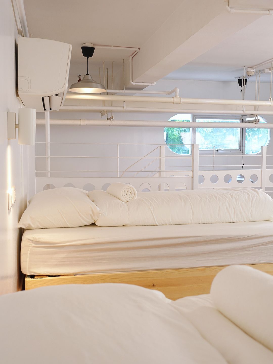 White bed in a room with white walls, pipes, and a window.
