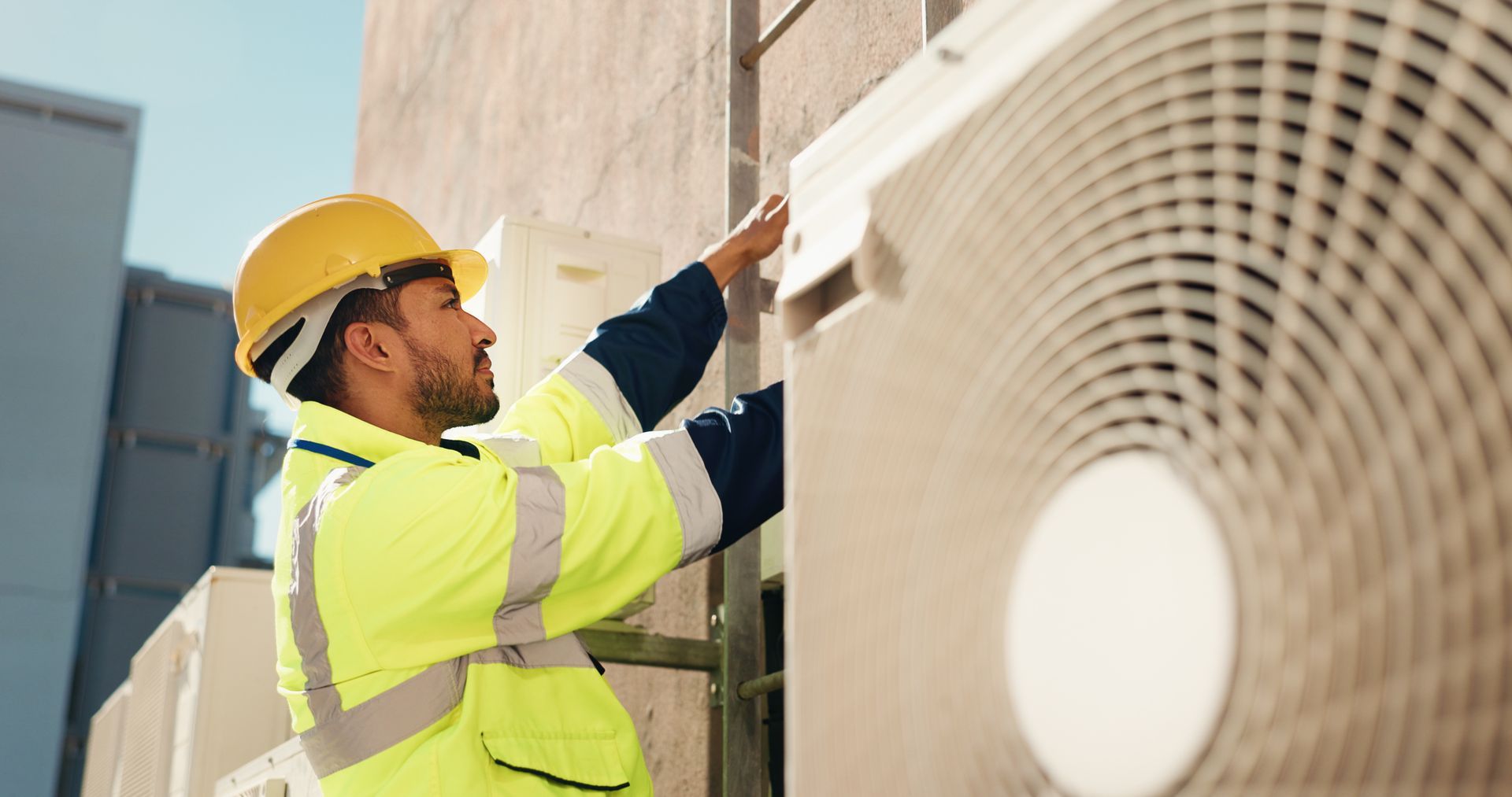 Technician in yellow safety gear inspecting an outdoor HVAC unit near a building wall
