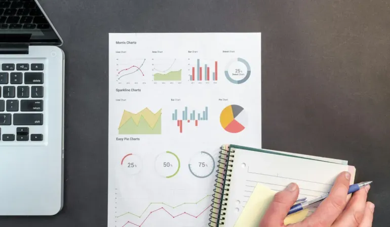 Laptop, charts, and notebook on a desk; a hand with pen is writing notes.