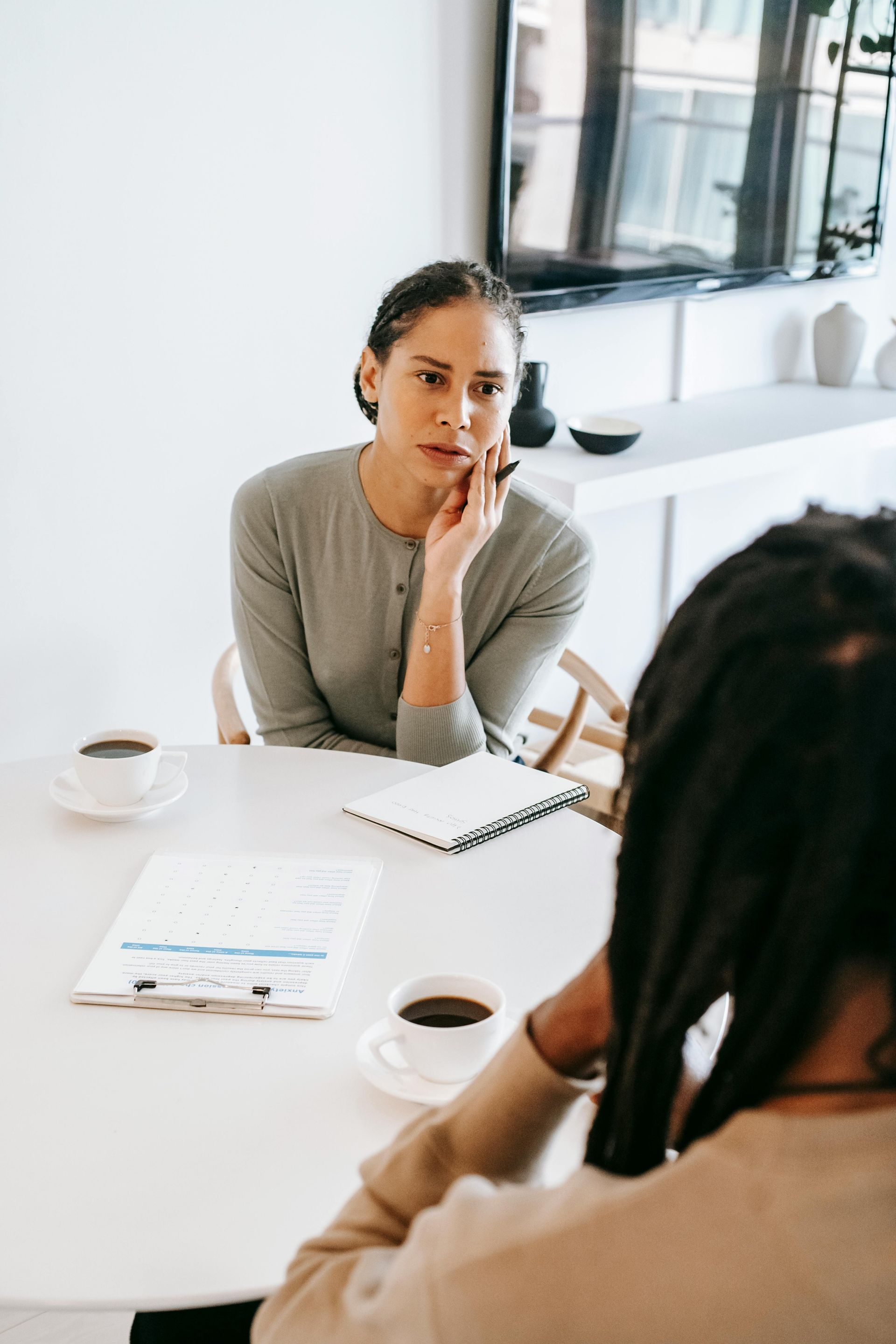 Woman seated at a table, hand to her face, listening intently to someone. Coffee and documents on the table.