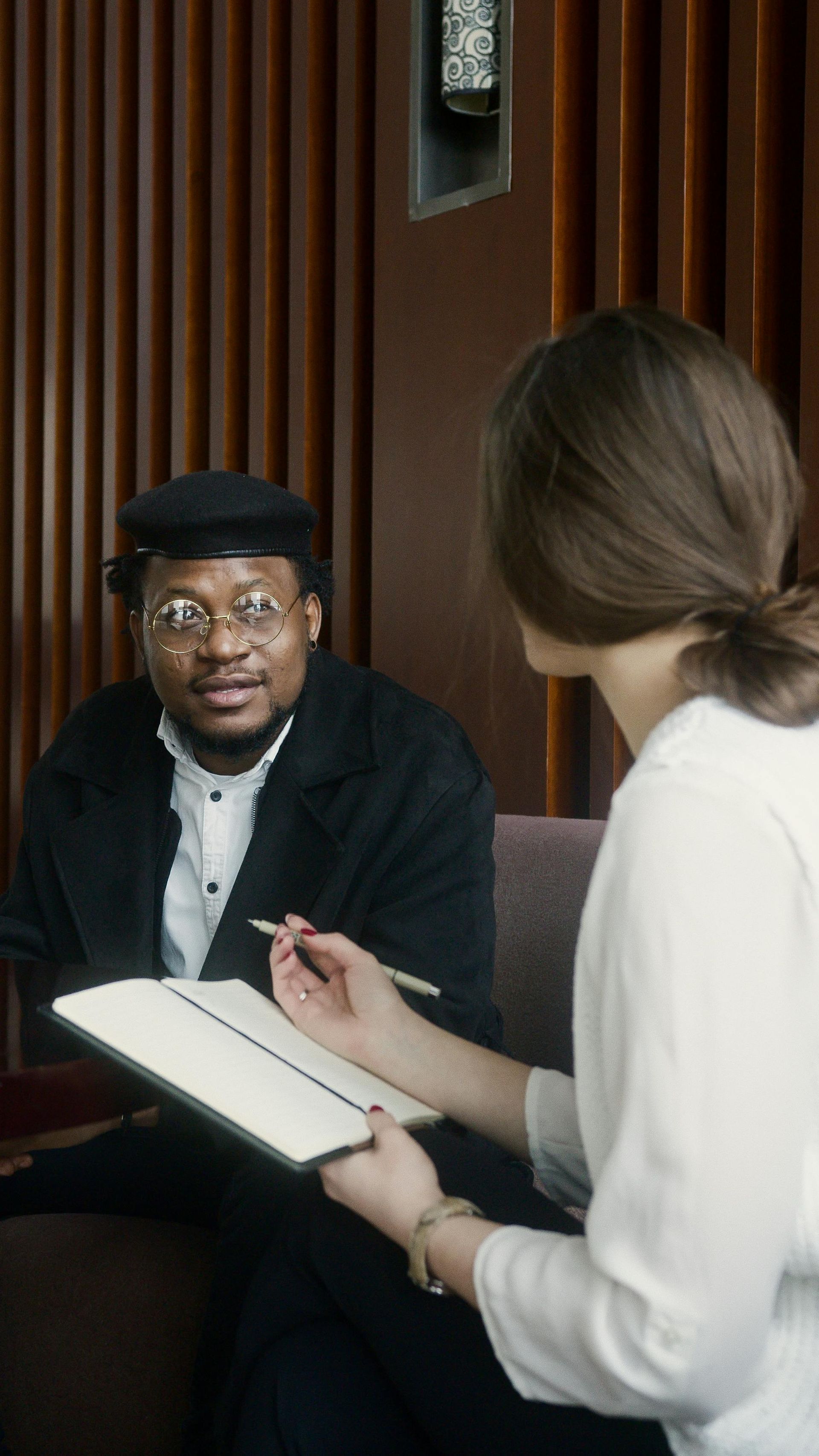 Man in conversation with person holding a notebook. Seated in a room with wood paneling.