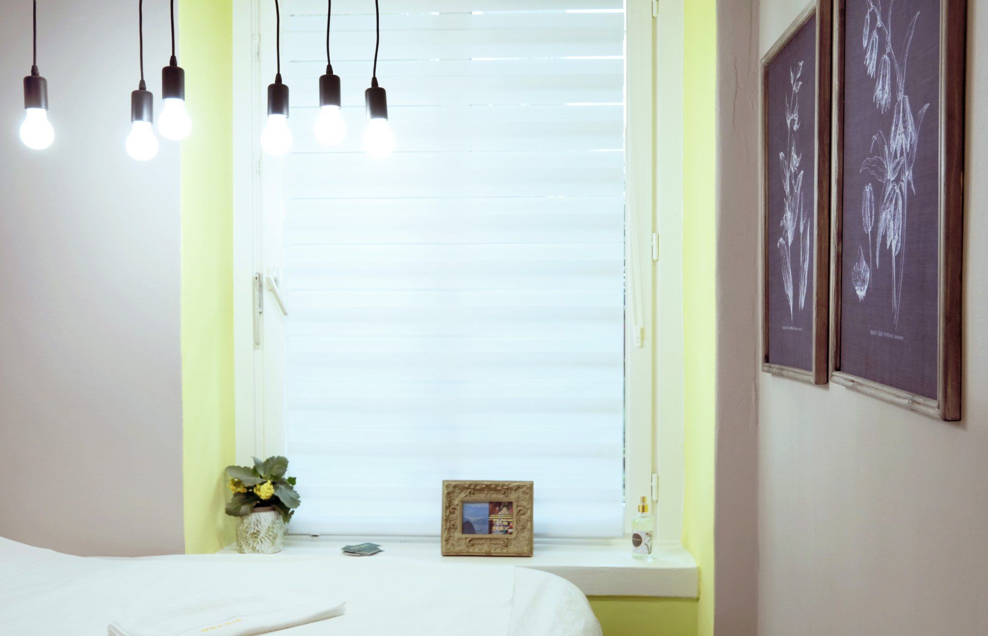 Schlafzimmer mit Fenster, Hängelampen, Kunstwerken, Bett und einem kleinen Bilderrahmen auf der Fensterbank.