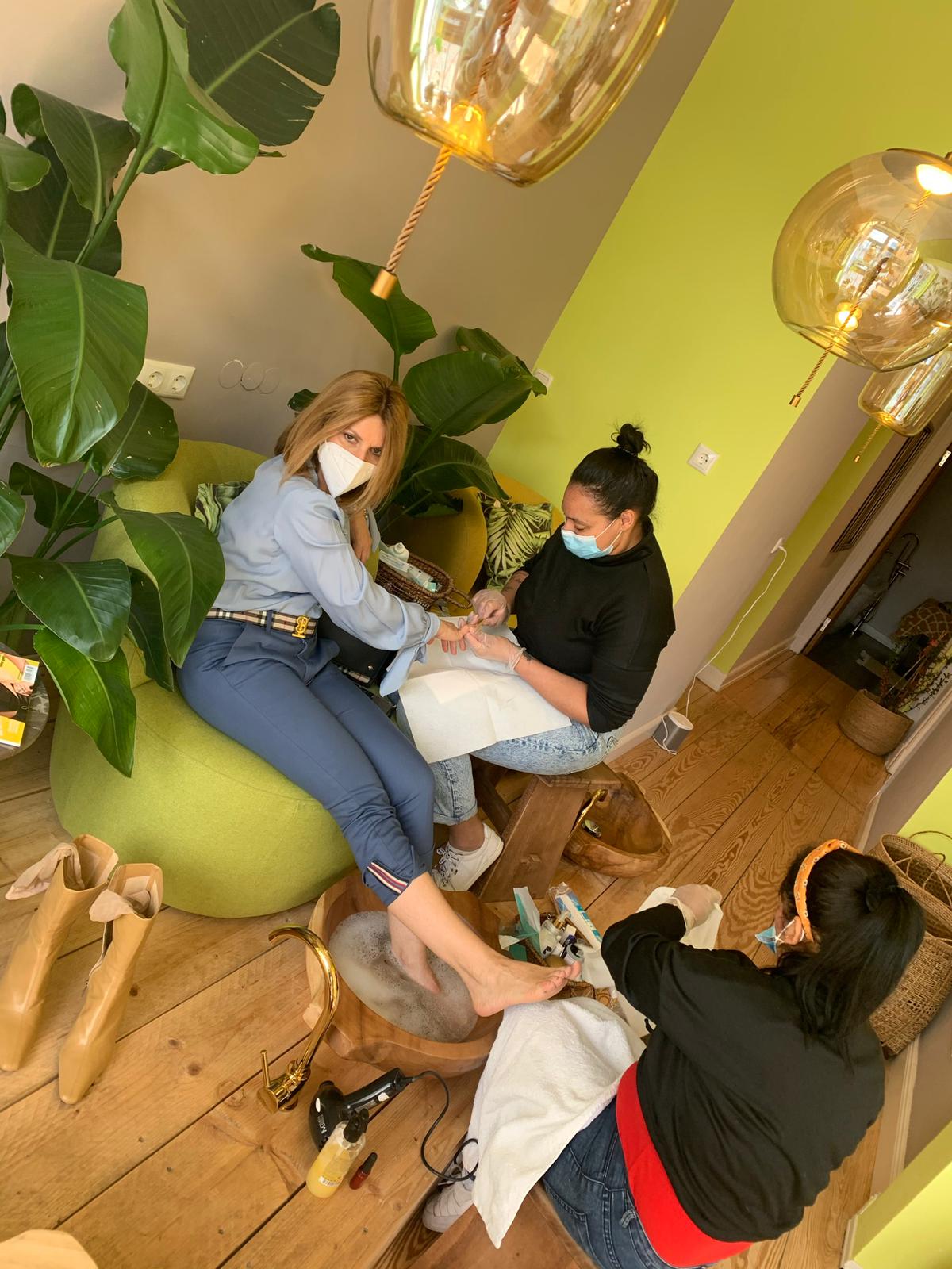 Eine Frau lässt sich in einem Salon die Nägel machen. Zwei Kosmetikerinnen mit Masken bedienen sie.
