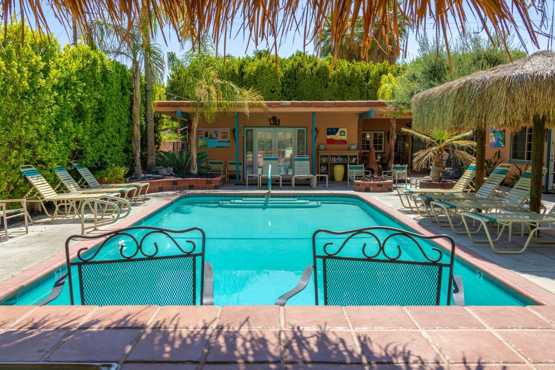 Swimming pool surrounded by lounge chairs and lush greenery under a thatched roof.