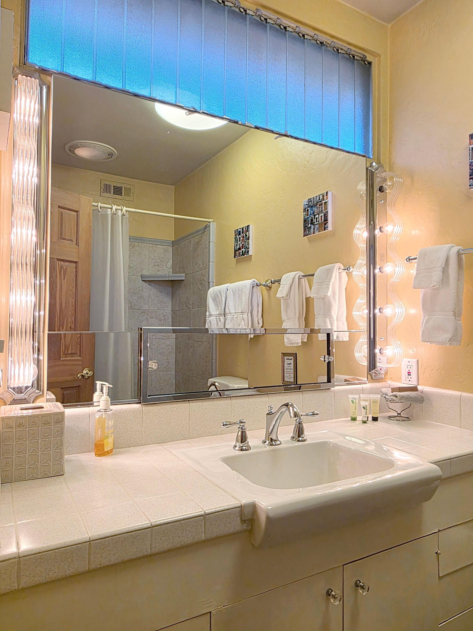Bathroom with a large mirror, sink, and light fixtures, reflecting a shower and towels.