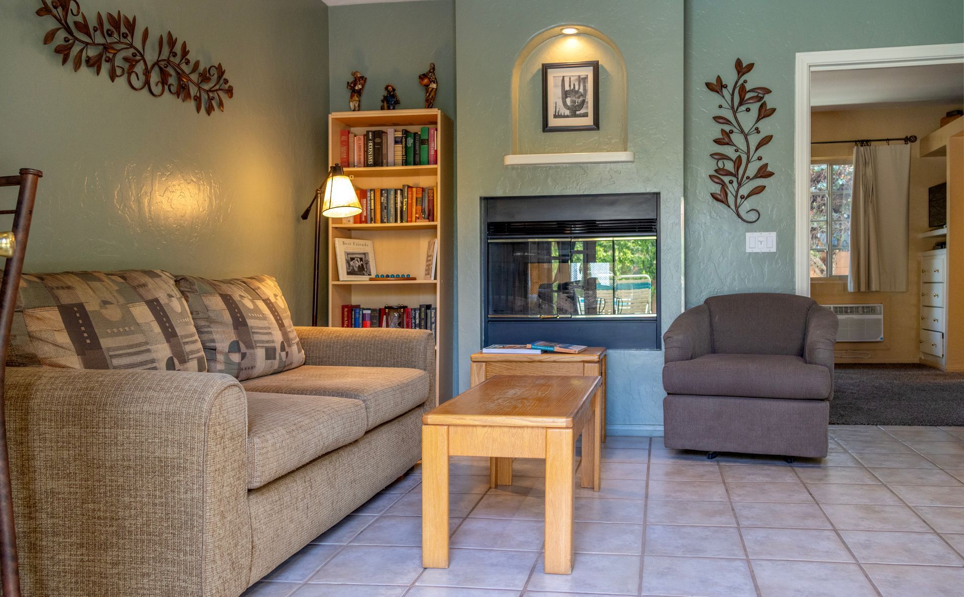 Cozy living room: tan couch, wooden bookshelf, fireplace, brown armchair, and a small wooden coffee table.