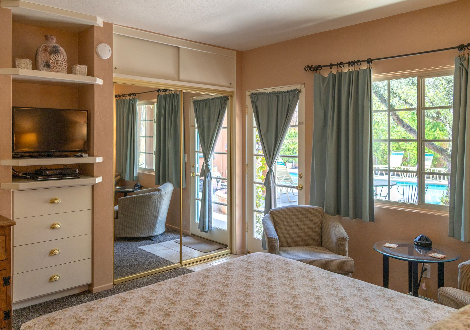 Bedroom with light peach walls, mirrored closet, window with blue curtains, and view of a pool.