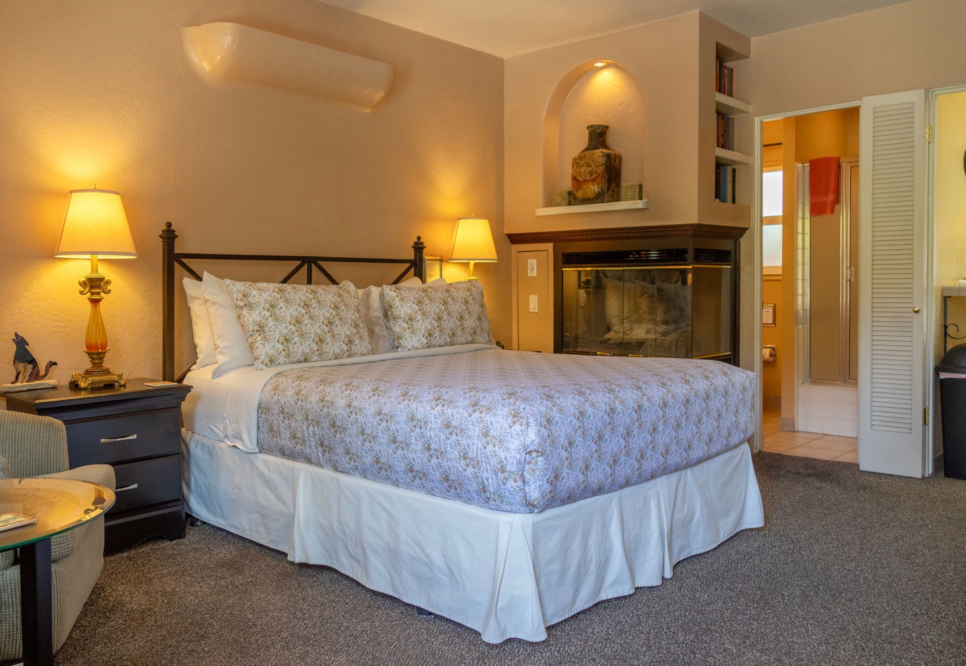 Cozy bedroom with a large bed, fireplace, lamps, and a bathroom visible in the background.