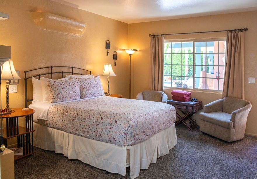 Bedroom with bed, two chairs, window with curtains, lamps, and small table.
