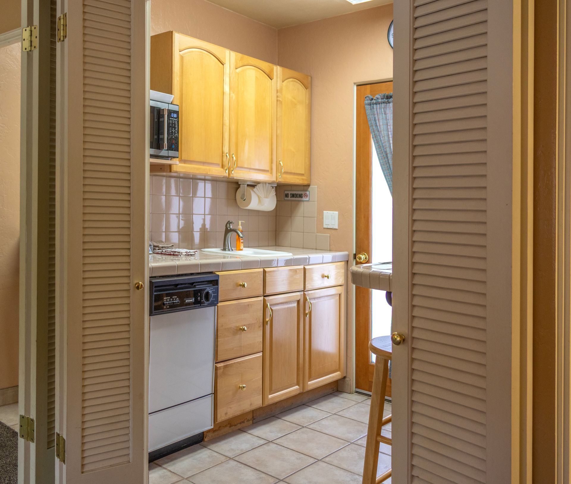 A small kitchen with light wood cabinets, a dishwasher, and a doorway with a counter and stool.