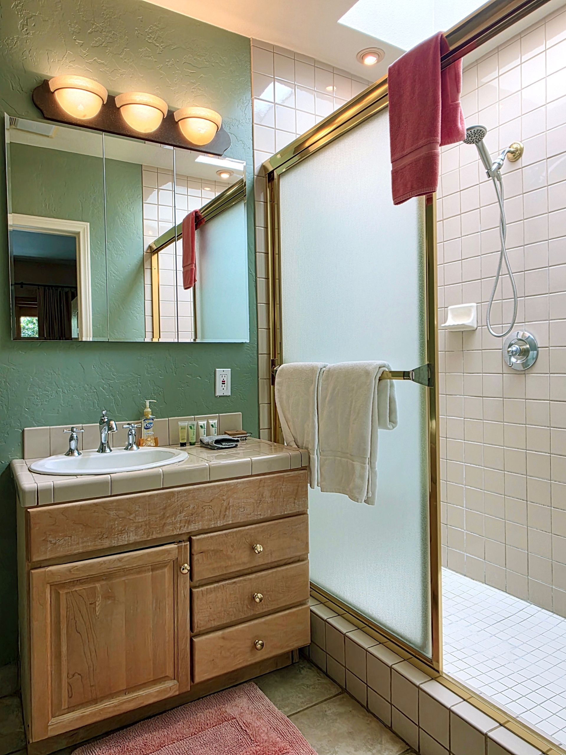 Bathroom with light-colored wood vanity, frosted glass shower, and green walls. Pink rug, white towels, and a skylight present.