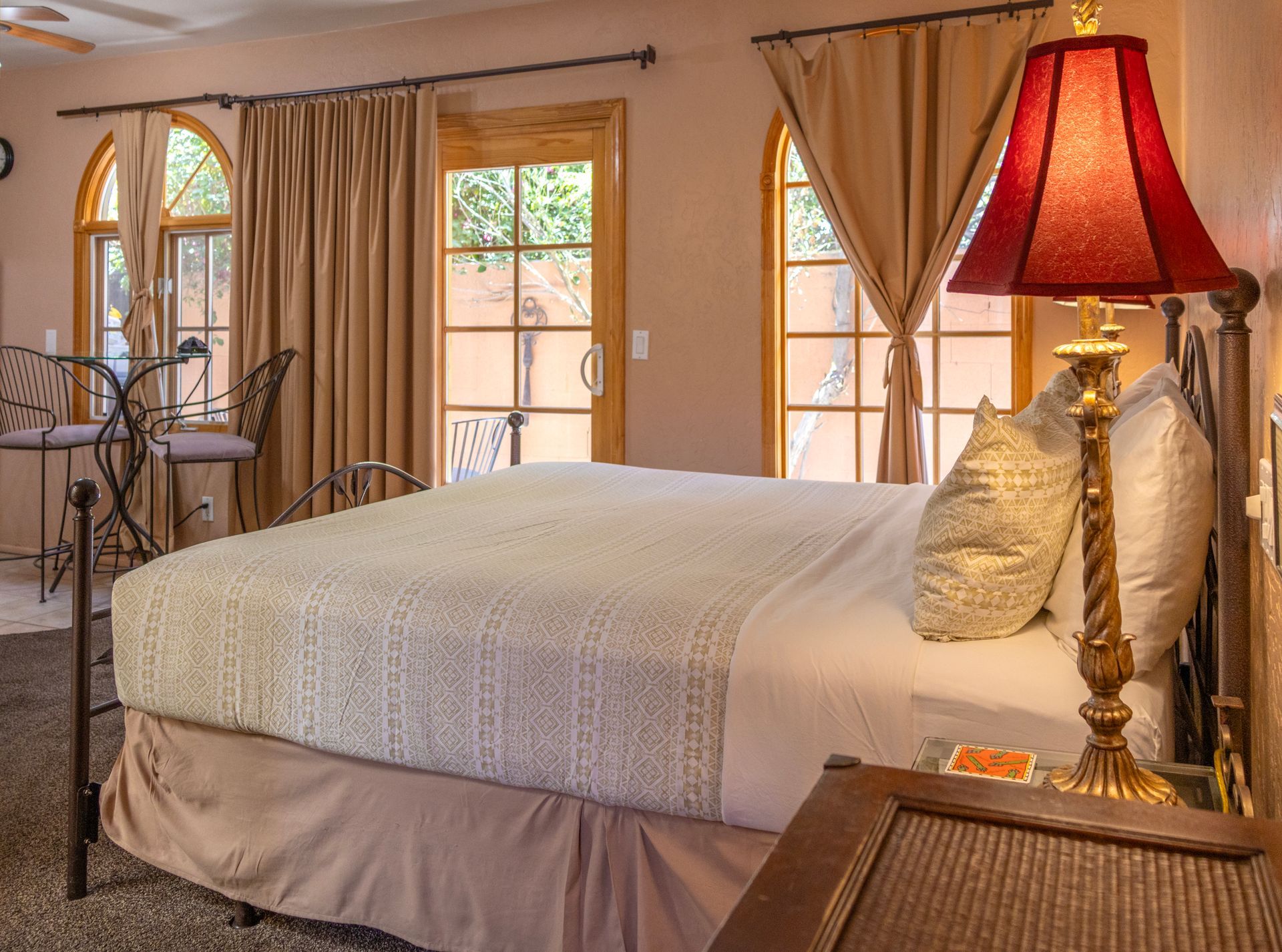 Bedroom with a bed, lamp, windows, and a small table. Earth tones, cozy atmosphere.