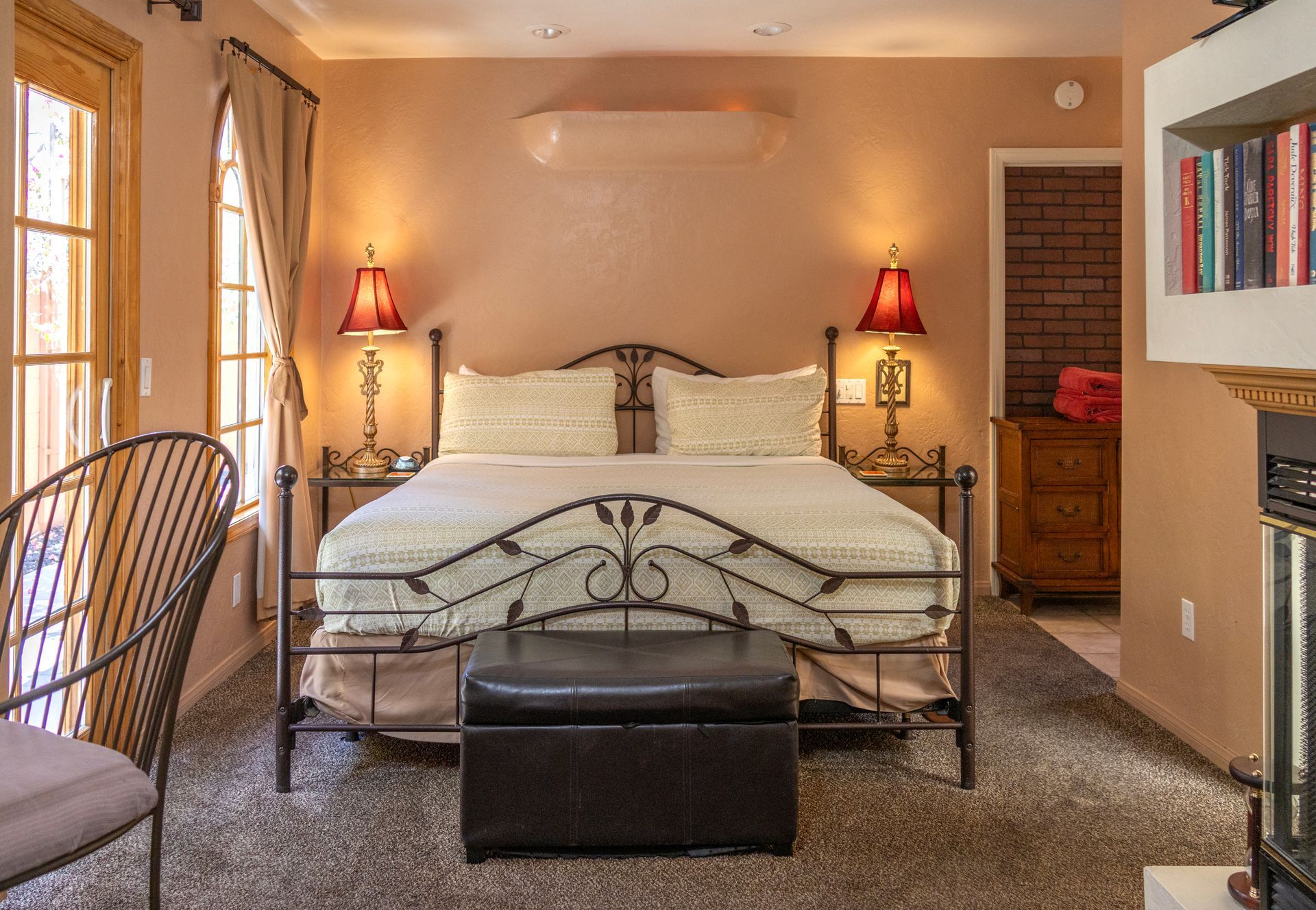 Bedroom with a wrought-iron bed, bedside lamps, and a leather ottoman. The room has a fireplace and a door to a balcony.