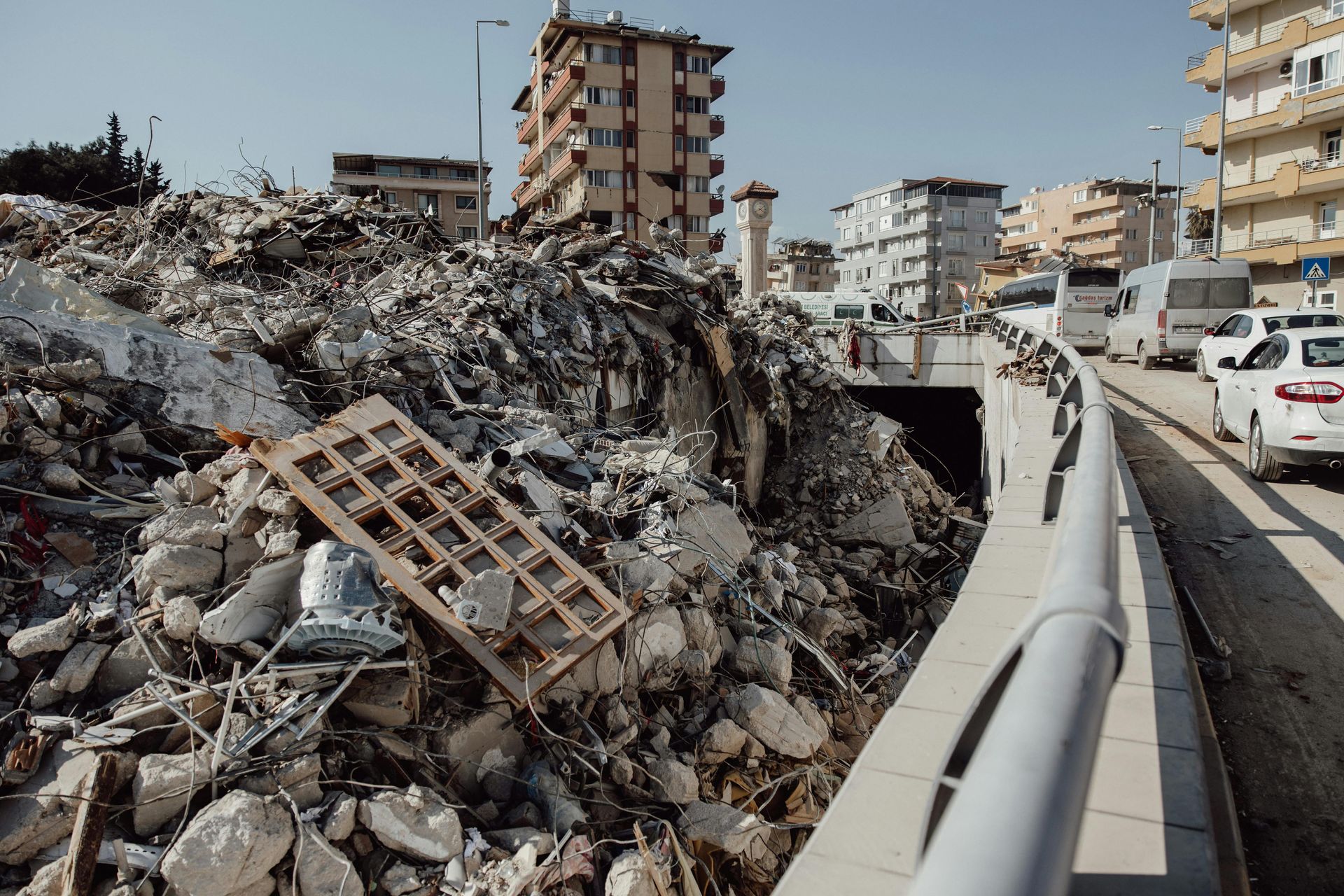 Debris pile from collapsed buildings on a city street with damaged buildings visible in the background; white vehicles drive on the side.