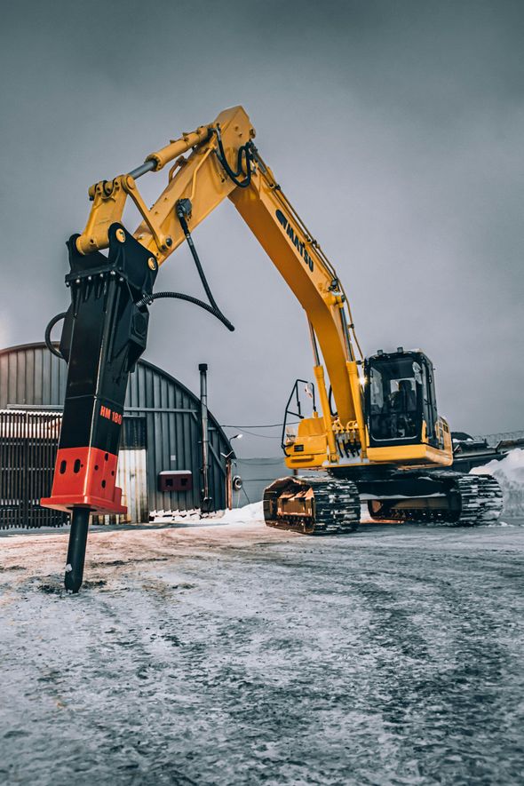 Yellow excavator with a jackhammer attachment, parked on snow-covered ground next to a building, under a cloudy sky.