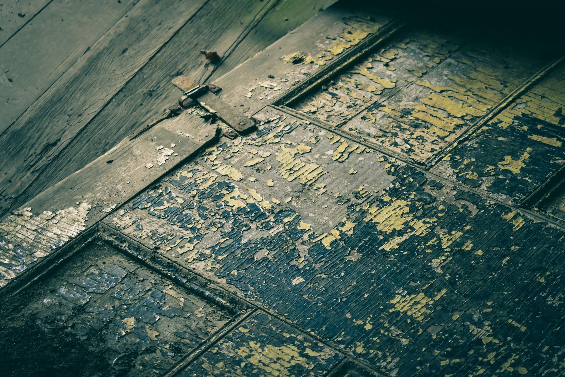 Close-up of weathered wooden floorboards with peeling blue and yellow paint. A metal hinge is visible.