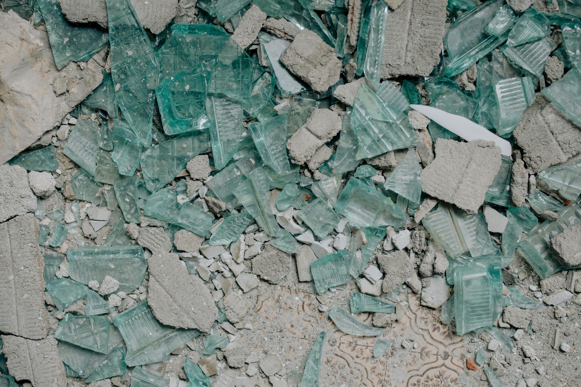 Broken shards of teal-colored glass and pieces of gray concrete debris scattered on a rough, light-colored surface.