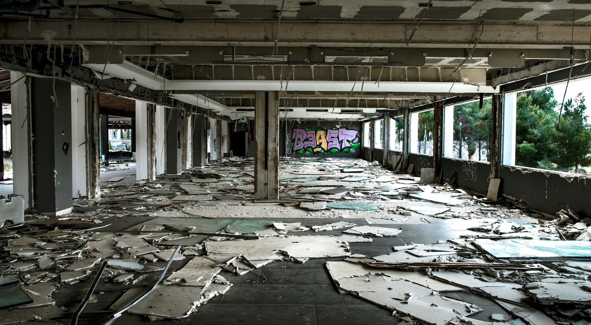 Interior of a ruined building with debris scattered across the floor, open to the outside with views of trees.