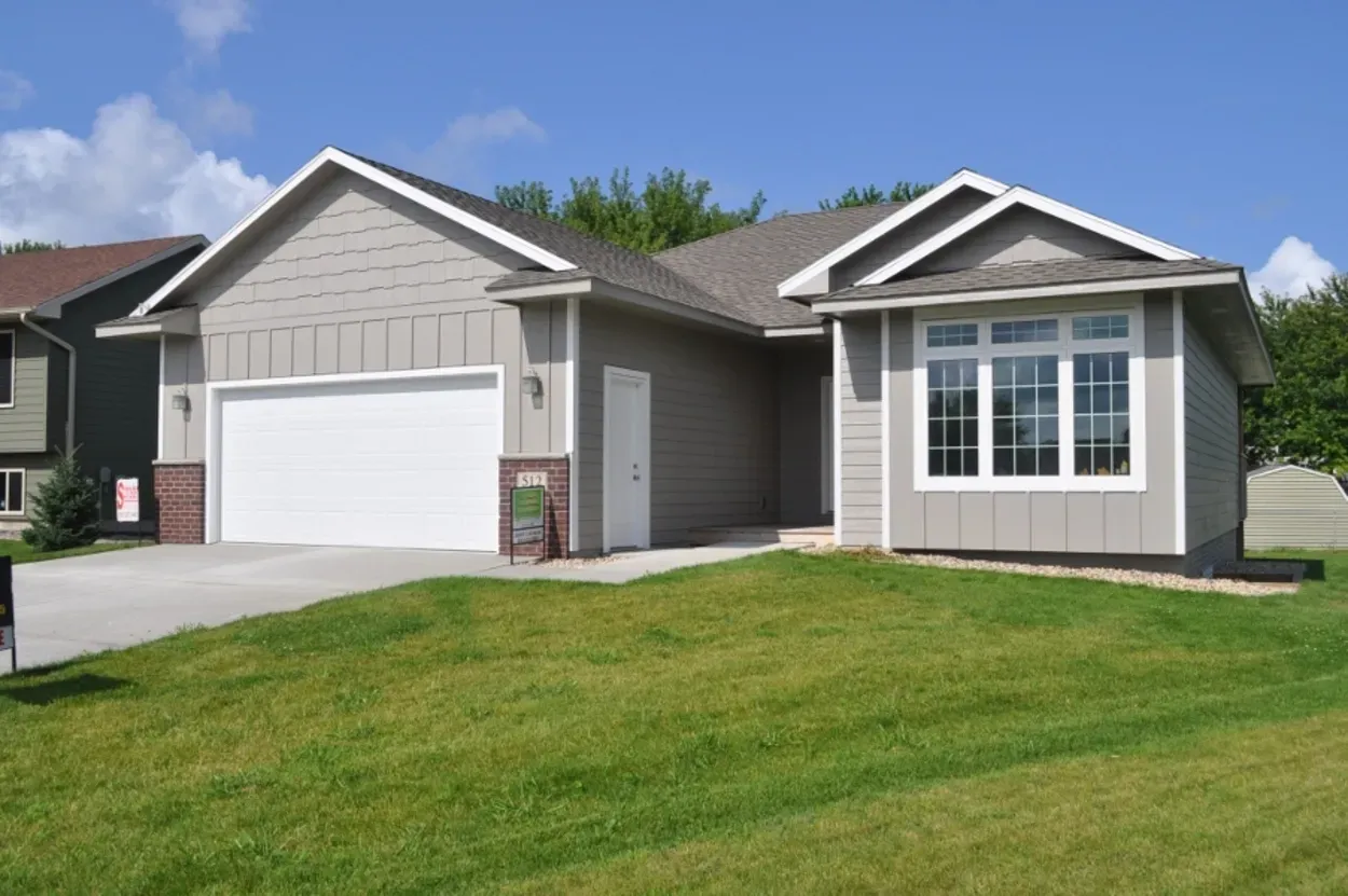Tan house with white garage door and bay window, green lawn, blue sky.