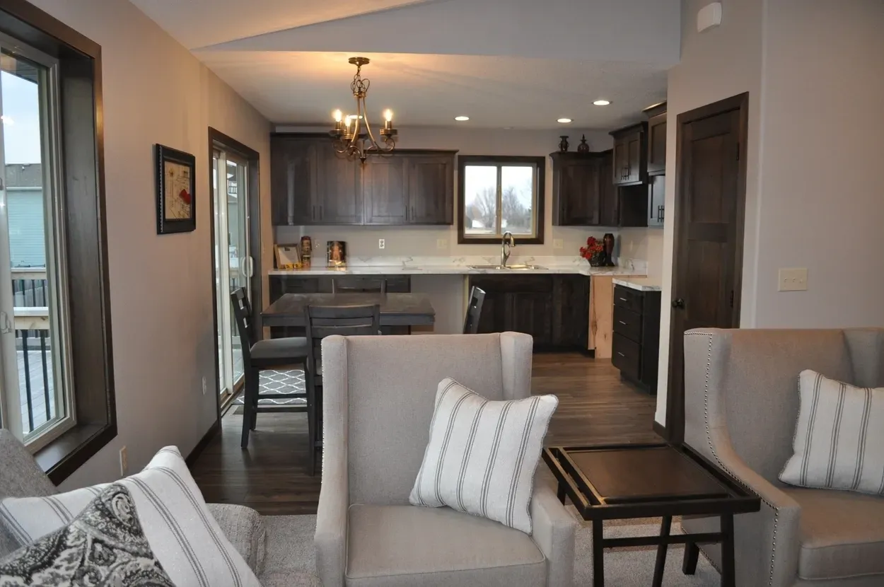 Cozy kitchen with dark cabinets, open to a living room with two chairs, neutral colors, and a chandelier.