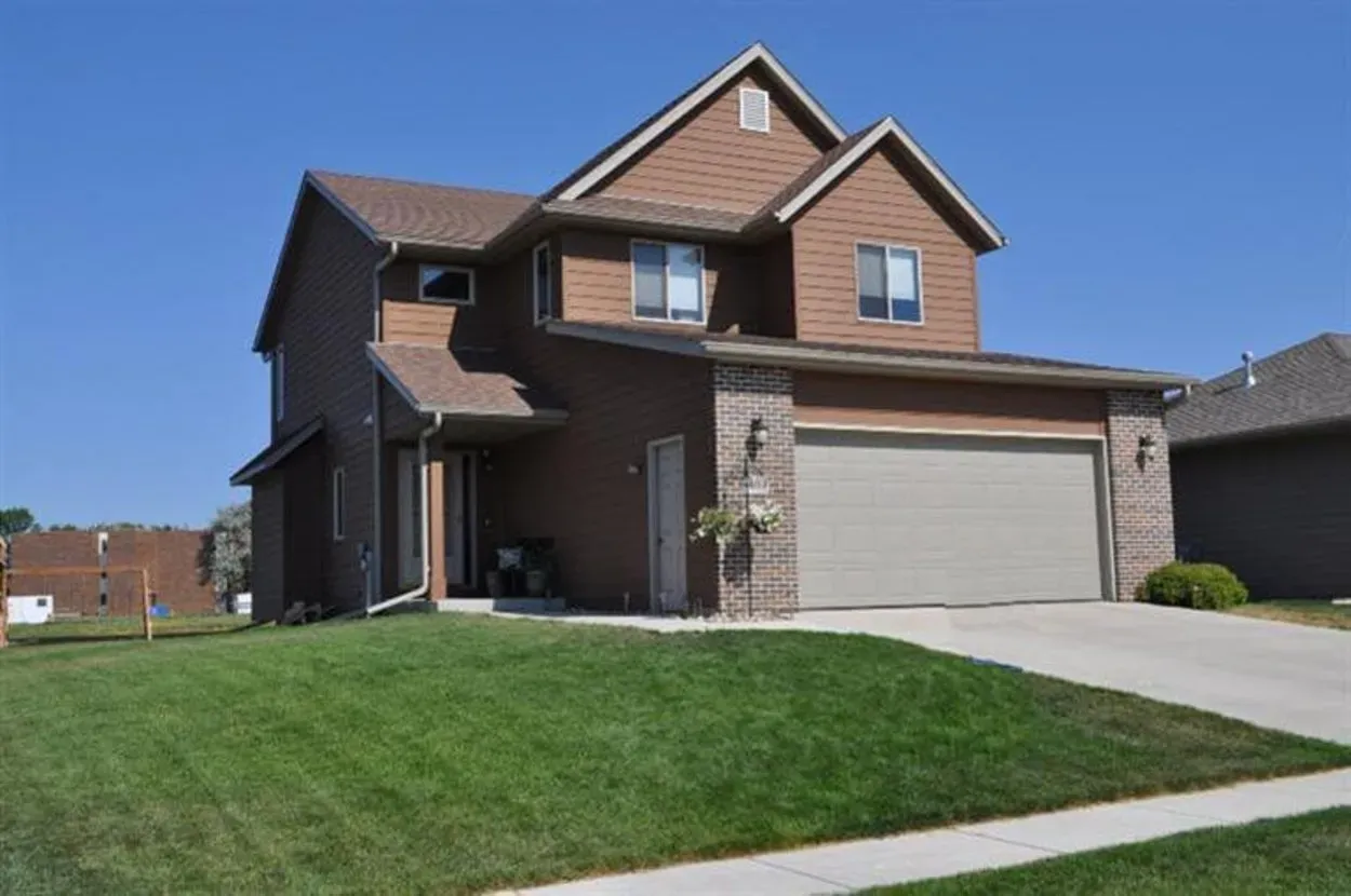 Two-story brown house with attached garage, beige door, and well-manicured lawn under a clear blue sky.