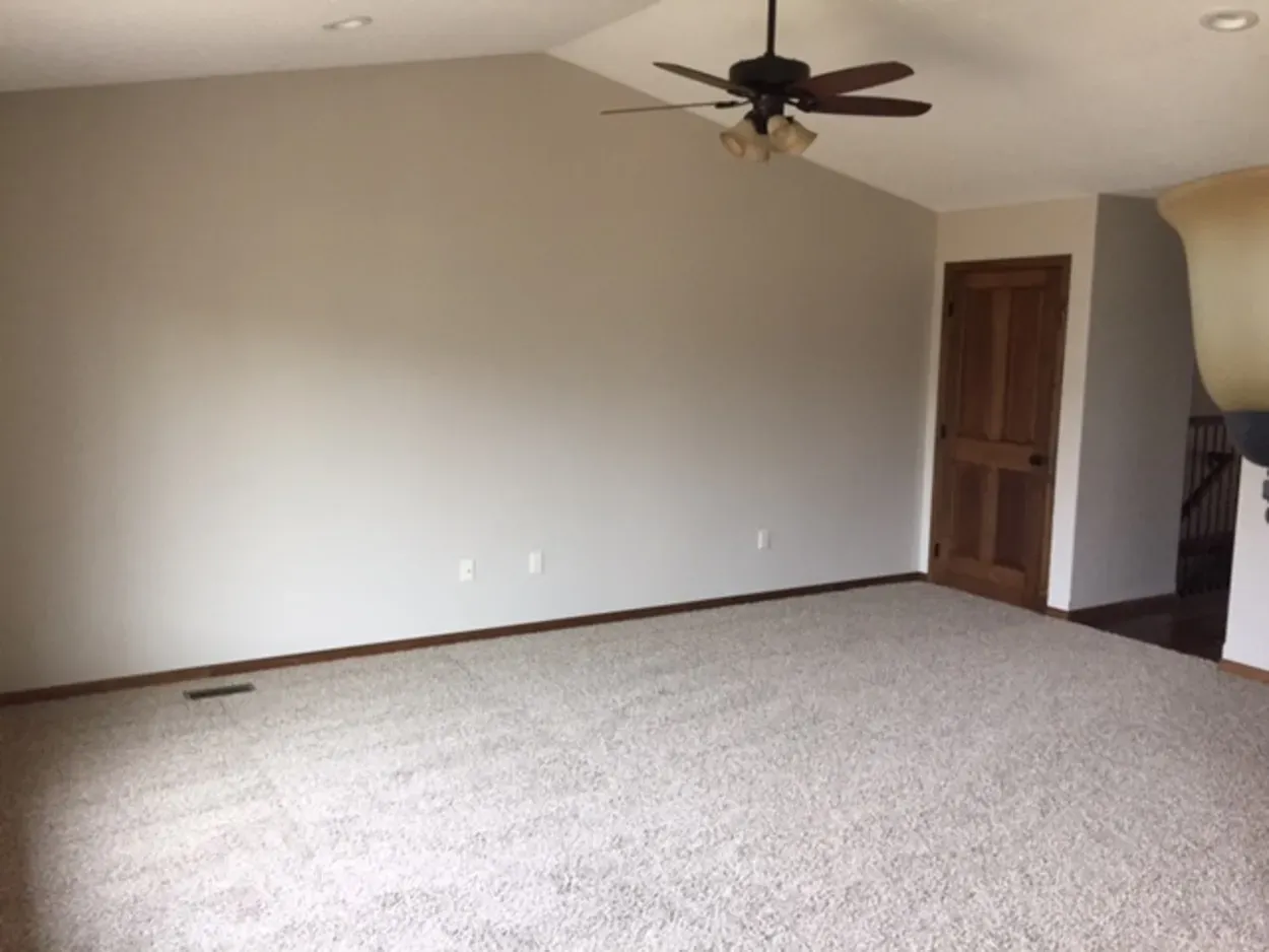 Empty room with beige walls, brown door, and beige carpet; a ceiling fan is visible.