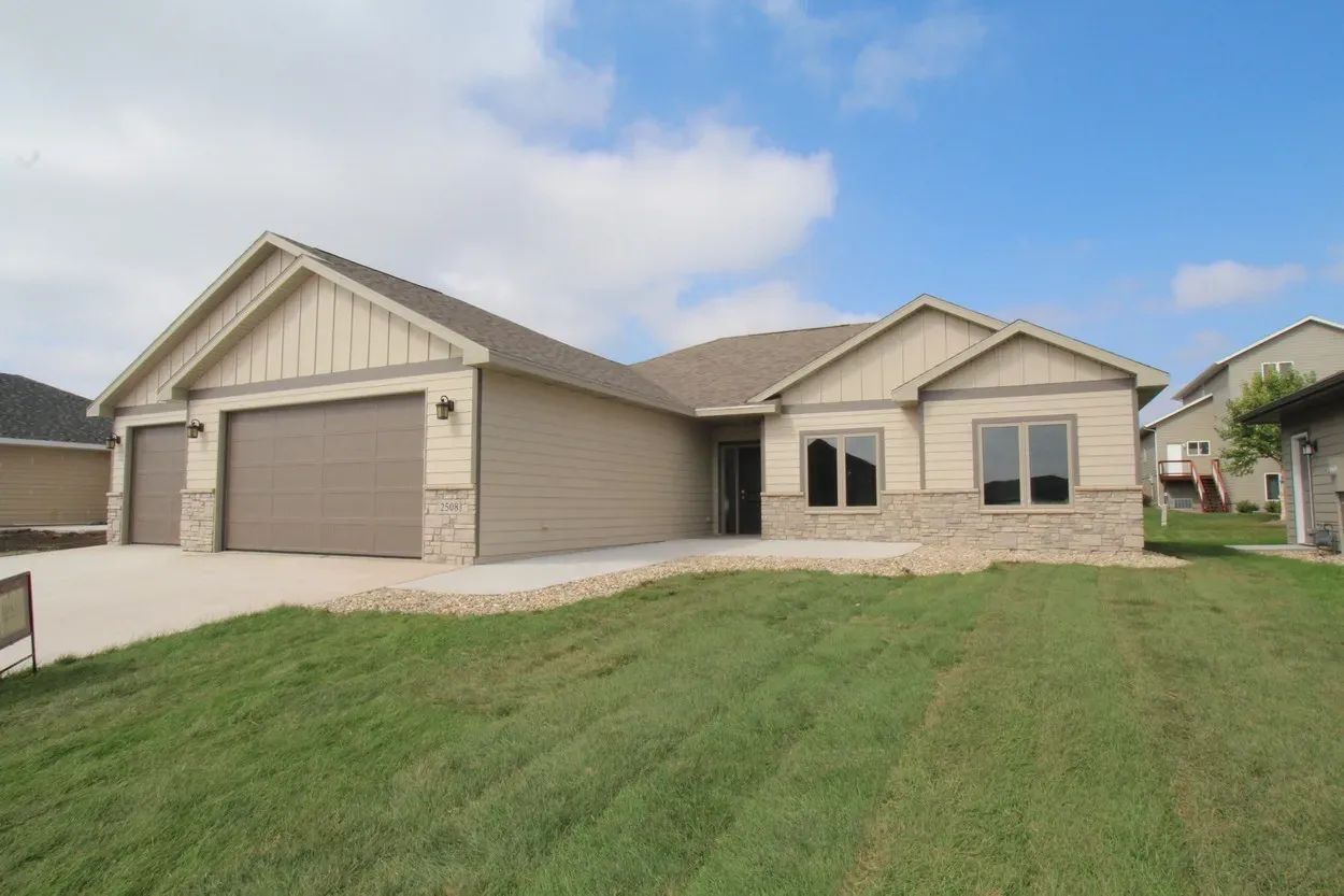 Beige ranch-style house with a two-car garage, brick accents, and a grassy lawn on a sunny day.