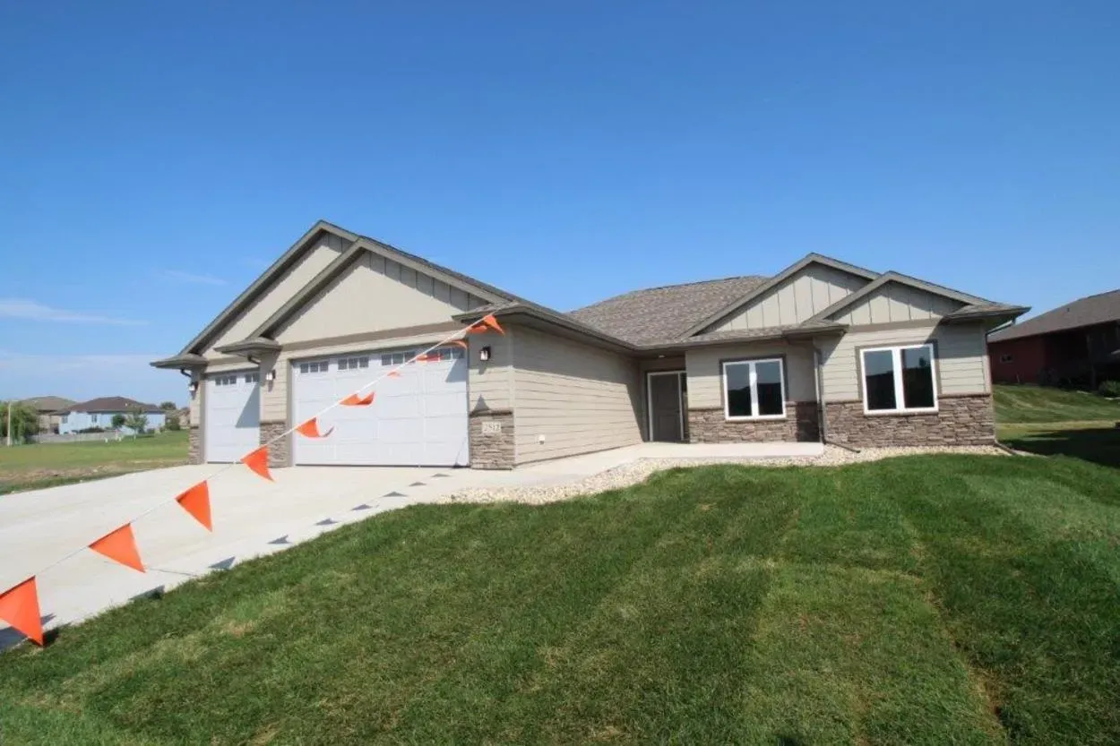 New beige house with a two-car garage, stone facade, and green lawn, under a blue sky.