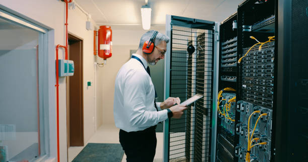 Man in blue jumpsuit working with computer servers, pointing at a cable.