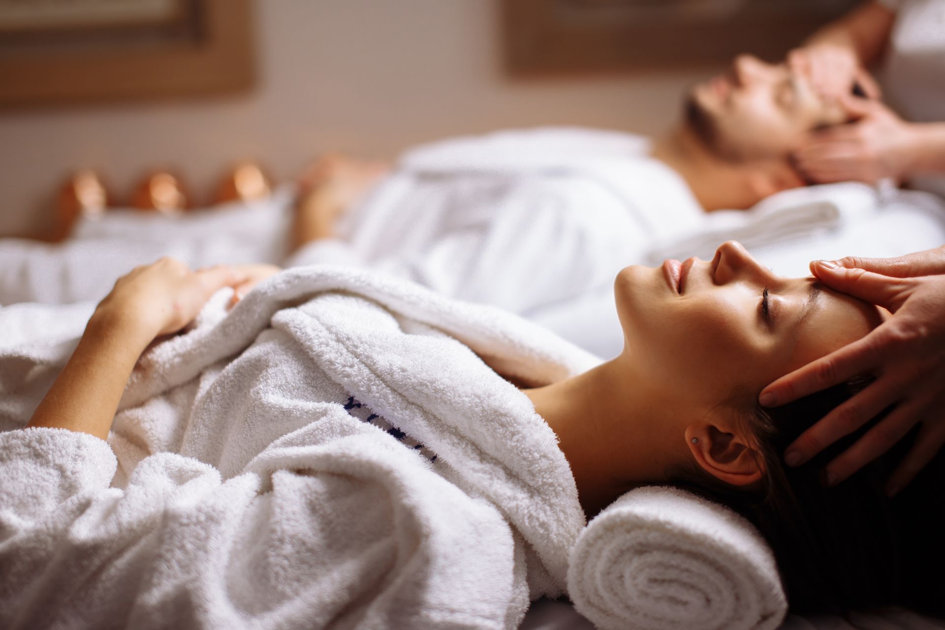 Two people in white robes receive head massages in a serene, dimly lit spa setting.
