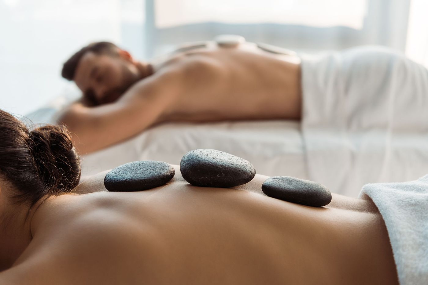 Two people lie on massage tables in a spa, receiving hot stone therapy with smooth dark stones placed on their backs.