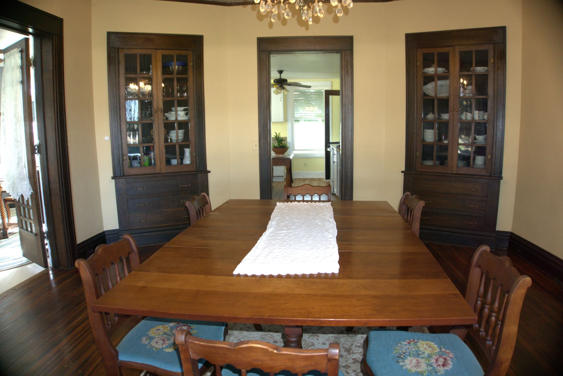 A dining room with a wooden table, chairs, and built-in cabinets. Sunlight shines through a doorway leading to a kitchen.