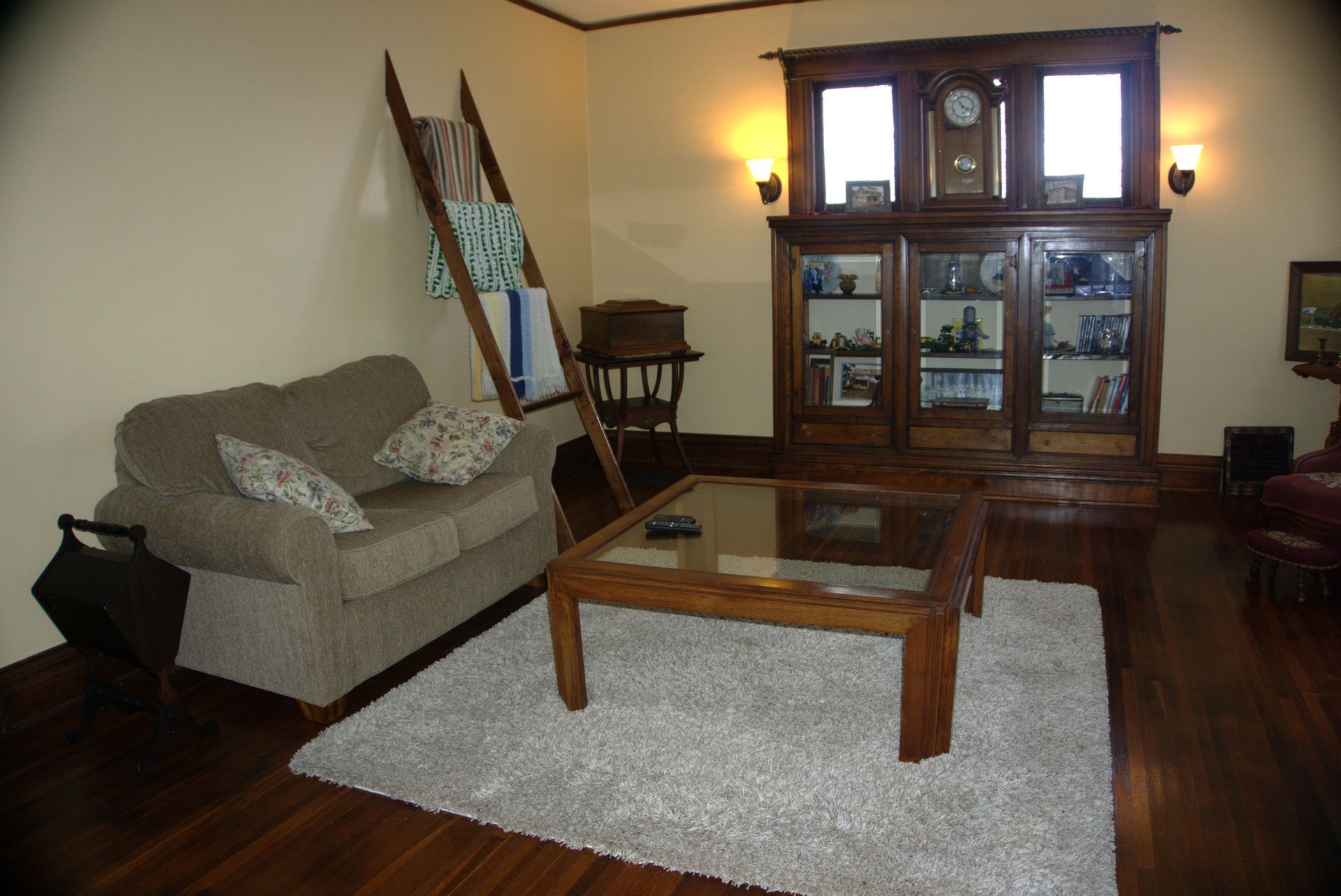 Living room with a tan couch, wooden coffee table on a white rug, and antique display cabinet. A ladder holds blankets.