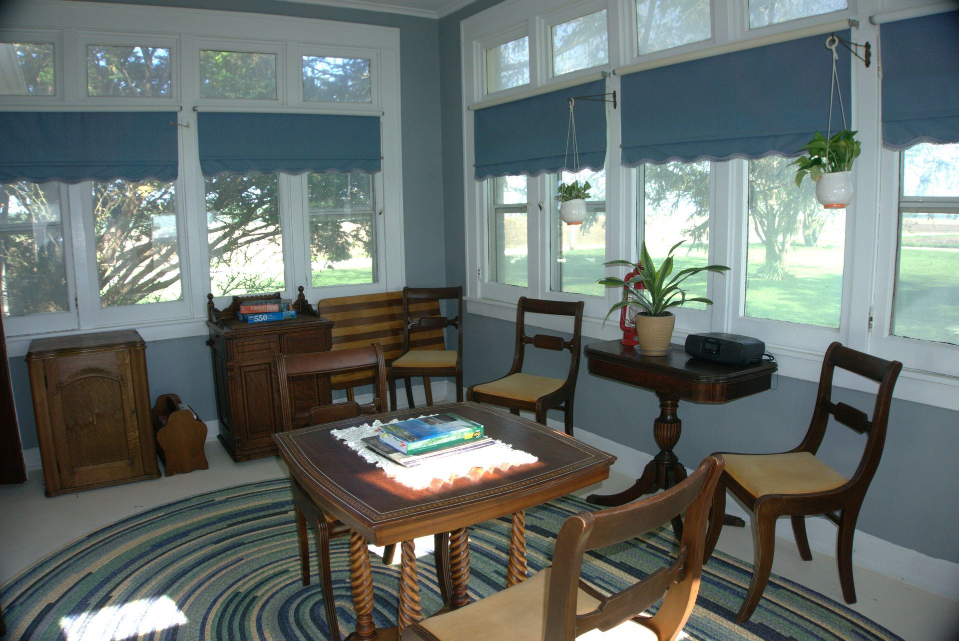 Sunroom with blue-gray walls and white-framed windows. Furnishings include wooden furniture, a blue rug, and potted plants, with a bright outdoor view.