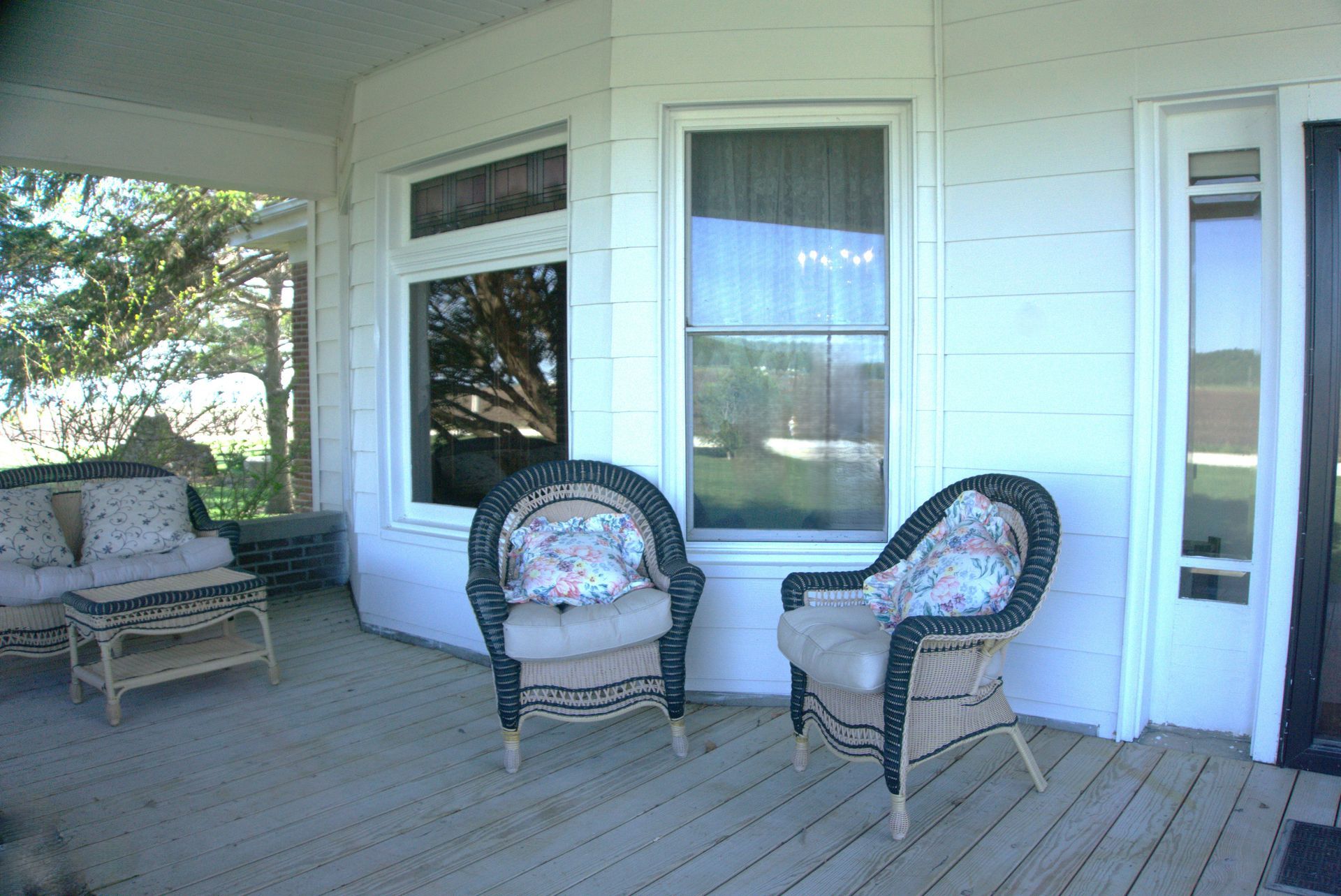 A porch with wicker furniture. Two chairs and a couch with floral cushions are on a wood-planked floor.