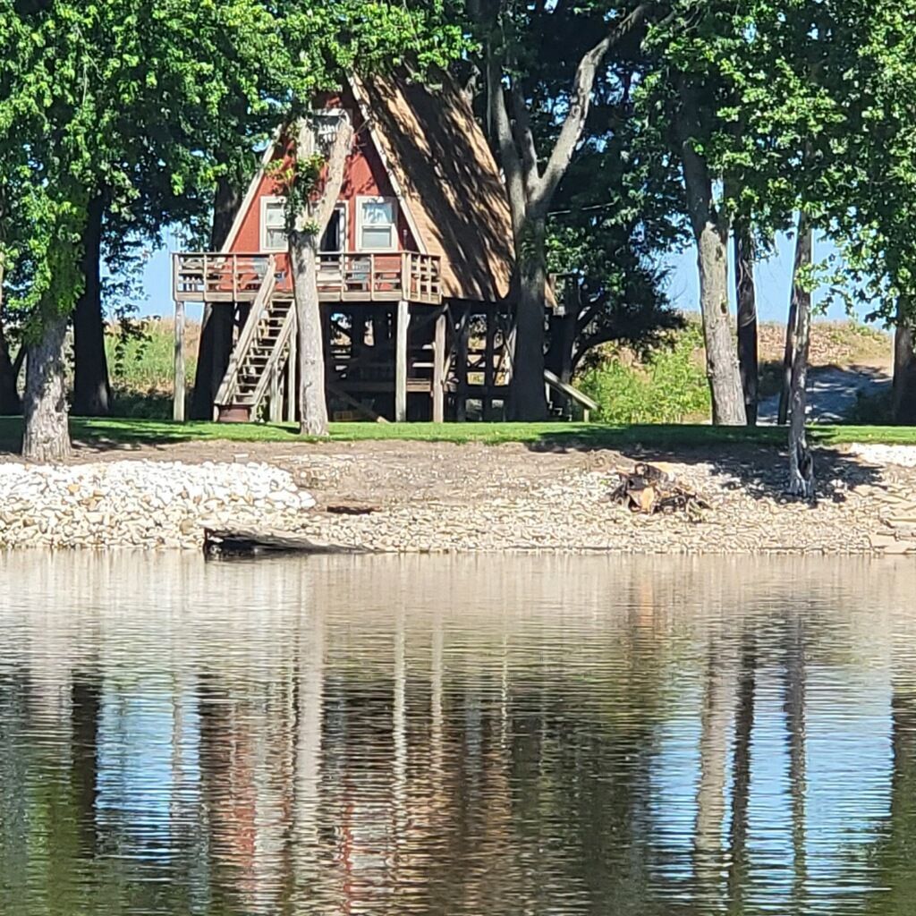 A-frame cabin on stilts with a red roof, nestled among green trees, reflected in the water.