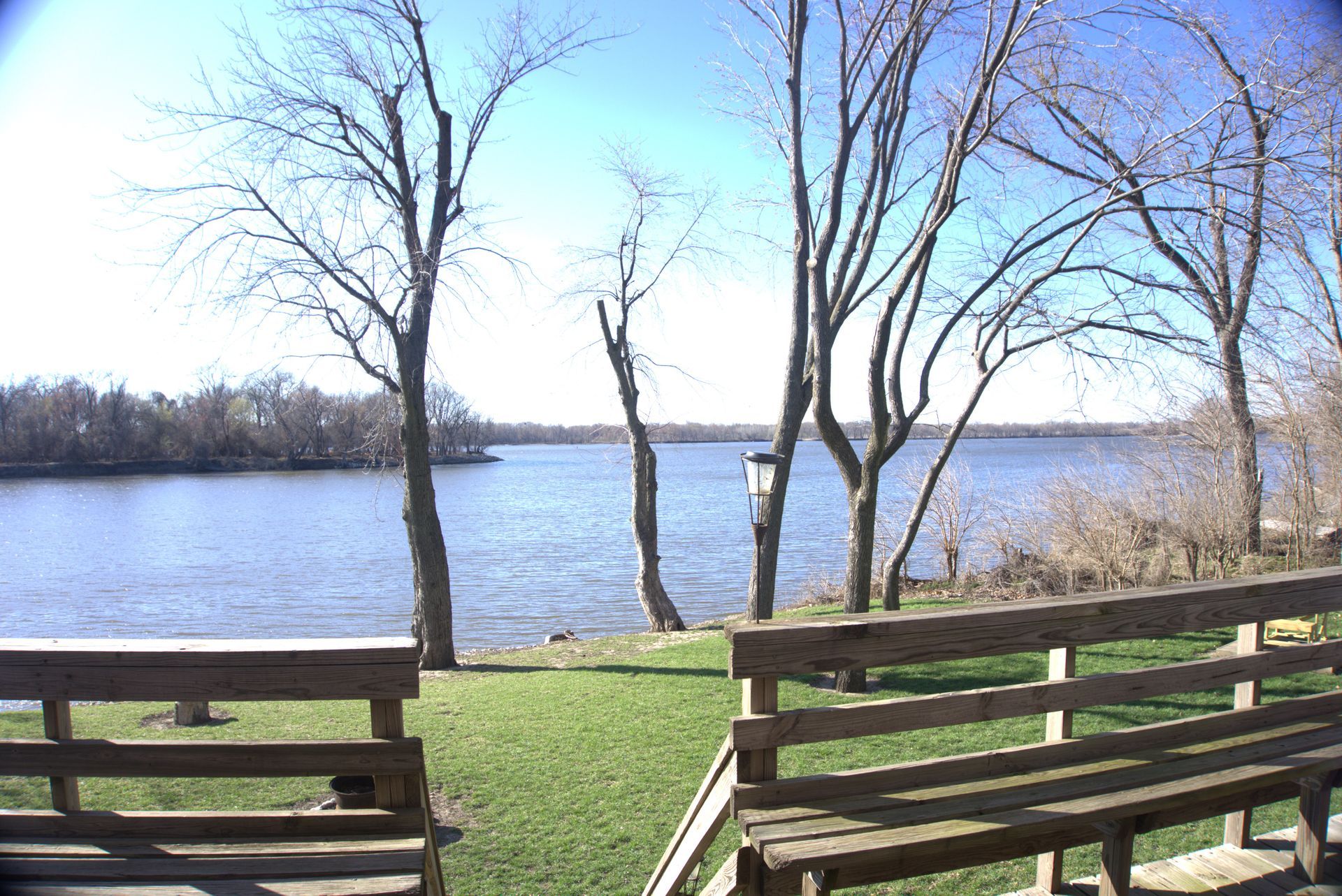 A view of a lake from a yard with a wooden fence. Several bare trees stand between the yard and the water.