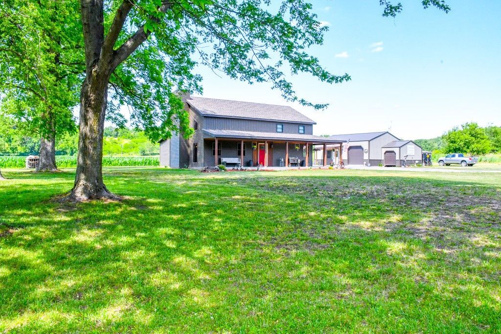A rustic house with a wrap-around porch and red door is set on a grassy lawn, shaded by a large tree.