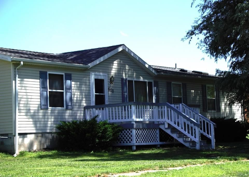A light-colored house with blue shutters and a white porch. A small lawn and a tree are in front of the house under a blue sky.