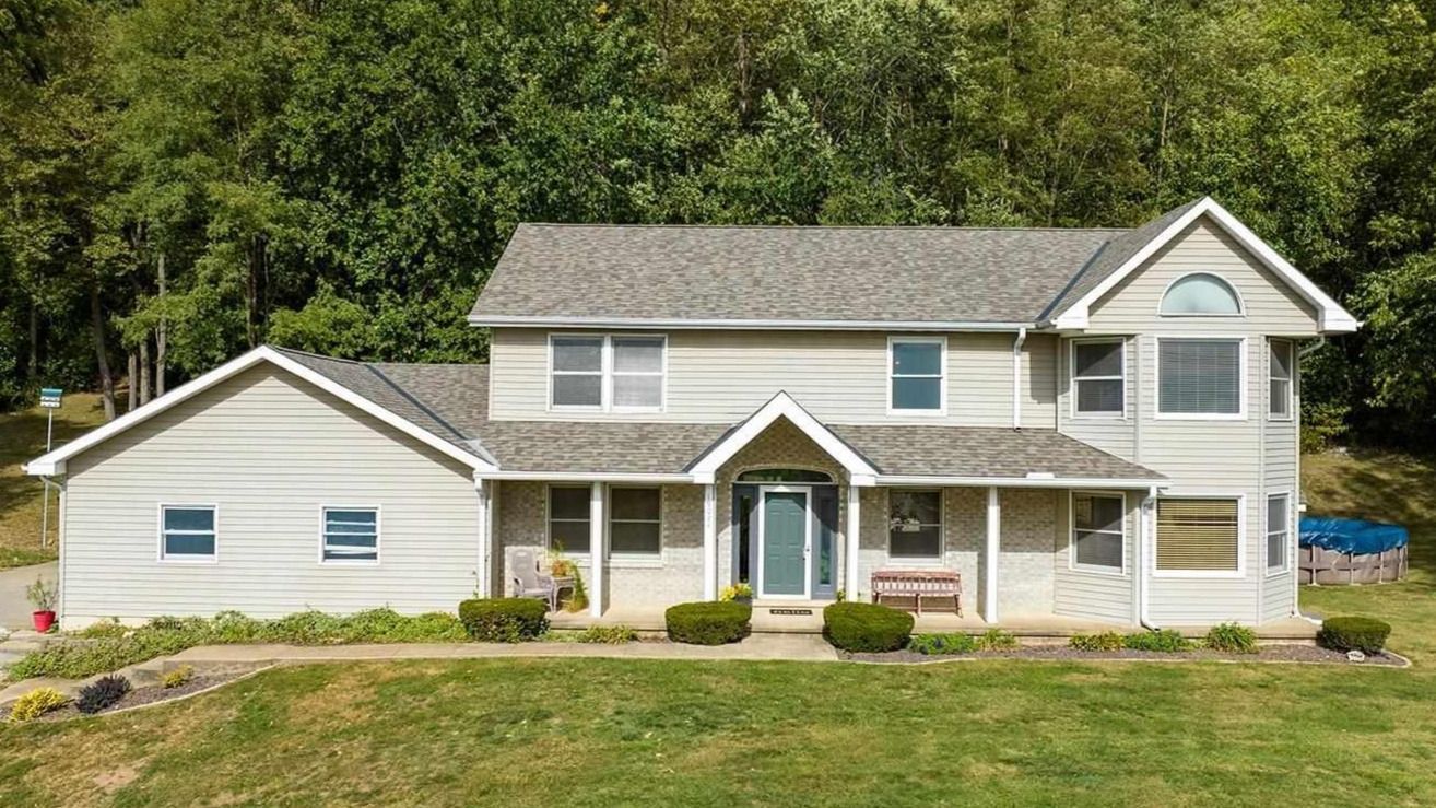 Two-story beige house with gray roof and front porch, set against a backdrop of green trees and grass.