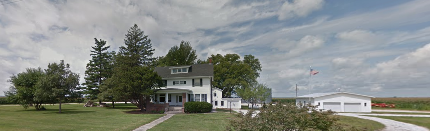 A large white farmhouse with a green lawn. A garage-like building is in the background under a cloudy sky.