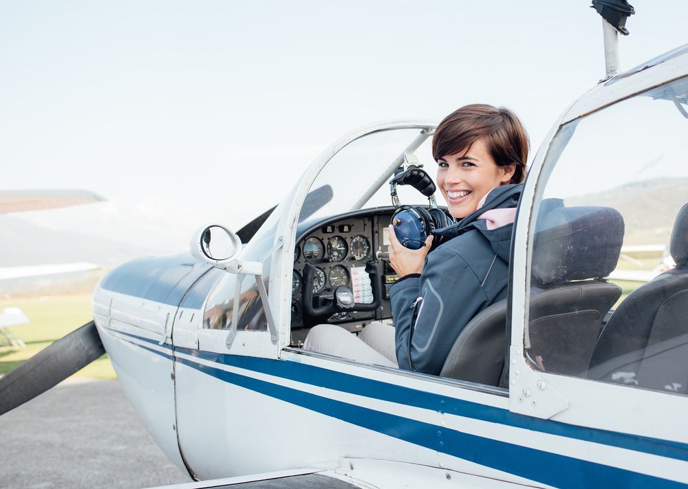 Smiling female pilot in the light aircraft cockpit, holding aviator headset and looking at camera.
