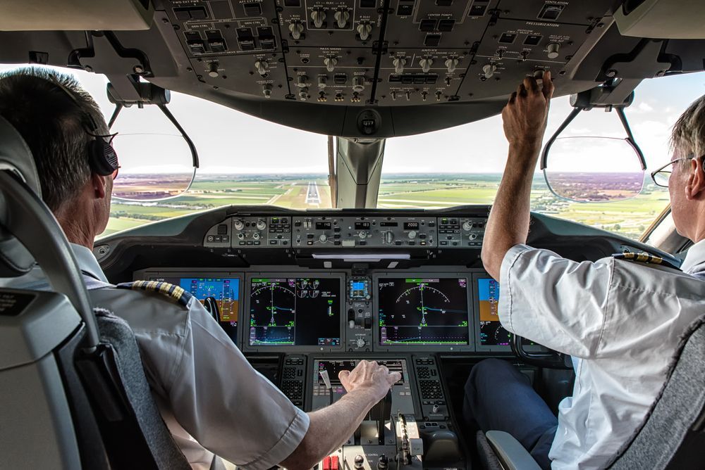 Pilots in cockpit, preparing to land. View shows instruments, runway, and pilot's hands on controls.