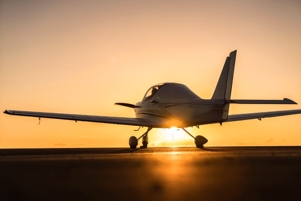 Small airplane on a runway, silhouetted by the setting sun, reflecting on the pavement.