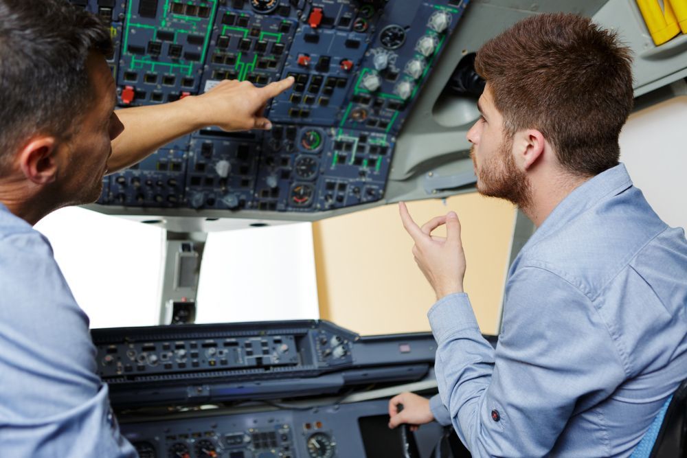 Two men are sitting in a cockpit of an airplane and one is pointing at something.